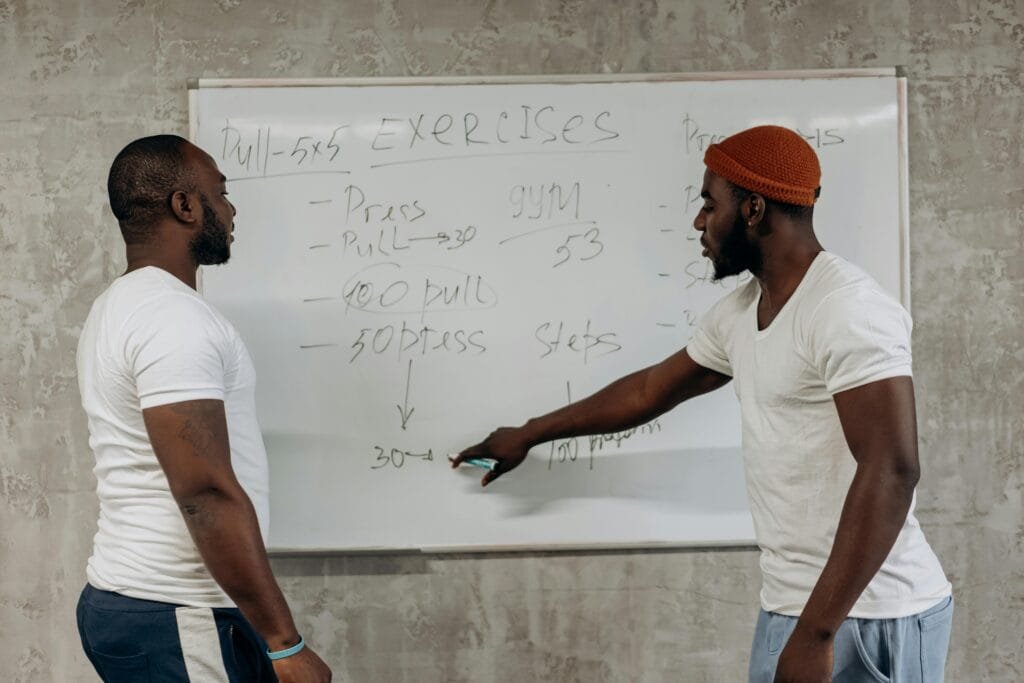 Two men discussing exercise routines on a whiteboard, planning gym workouts indoors.