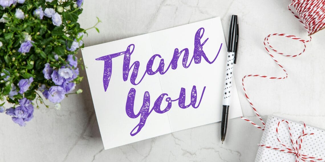 A stylish thank you card paired with purple flowers, a gift box, and pen on a marble surface.