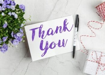A stylish thank you card paired with purple flowers, a gift box, and pen on a marble surface.