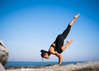 Woman performing a yoga pose on a rocky beach with ocean view and clear blue sky.