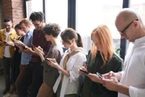 Diverse group of adults interacting with smartphones indoors by a large window, engaged in digital communication.