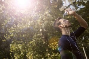 A man pours water over his head to cool off on a sunny day in a leafy outdoor setting.