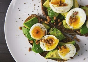 Delicious avocado toast topped with soft-boiled eggs and pine nuts on a white plate.