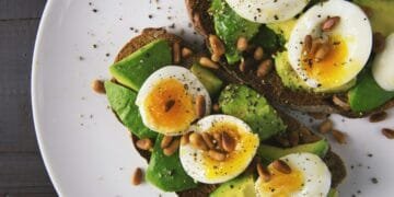 Delicious avocado toast topped with soft-boiled eggs and pine nuts on a white plate.