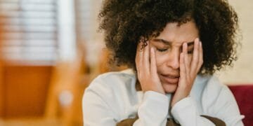 Despaired African American female with closed eyes touching face while sitting with pillow in light room at home on blurred background