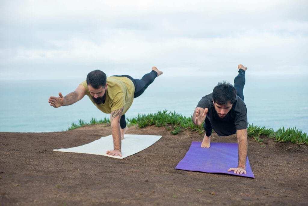 Two men practicing yoga poses on mats by the ocean, promoting fitness and well-being.