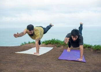 Two men practicing yoga poses on mats by the ocean, promoting fitness and well-being.