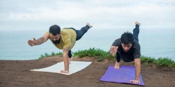 Two men practicing yoga poses on mats by the ocean, promoting fitness and well-being.