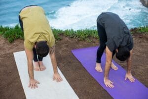 Two men practicing yoga outdoors by the sea in Portugal, performing forward bends on yoga mats.