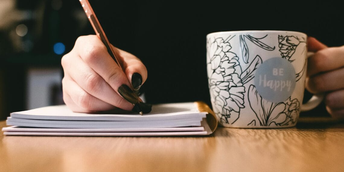 Person writing in a notebook with a floral ceramic mug on a wooden desk.