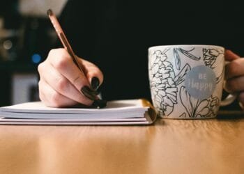 Person writing in a notebook with a floral ceramic mug on a wooden desk.