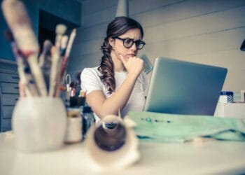 Young woman with glasses deeply focused on a laptop surrounded by art supplies in a home office.