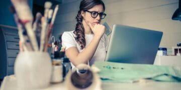Young woman with glasses deeply focused on a laptop surrounded by art supplies in a home office.