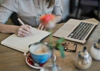 Woman writing in a notebook with a laptop and coffee cup on a desk. Ideal for workspace inspiration.