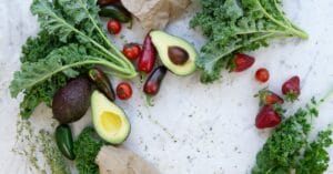 Top view of fresh avocados, kale, tomatoes, and peppers arranged on a marble surface.