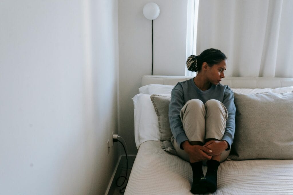 A young woman sits alone on a bed, looking upset and lonely in a cozy room.