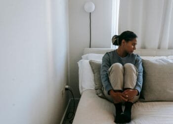 A young woman sits alone on a bed, looking upset and lonely in a cozy room.