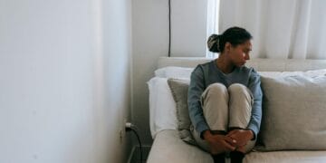 A young woman sits alone on a bed, looking upset and lonely in a cozy room.