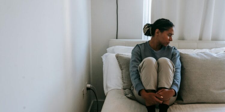 A young woman sits alone on a bed, looking upset and lonely in a cozy room.