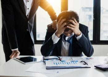 Professional feeling exhausted and wired under stressful workload. An overwhelmed businessman grasps his head, surrounded by reports in a bright office.