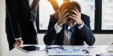 Professional feeling exhausted and wired under stressful workload. An overwhelmed businessman grasps his head, surrounded by reports in a bright office.