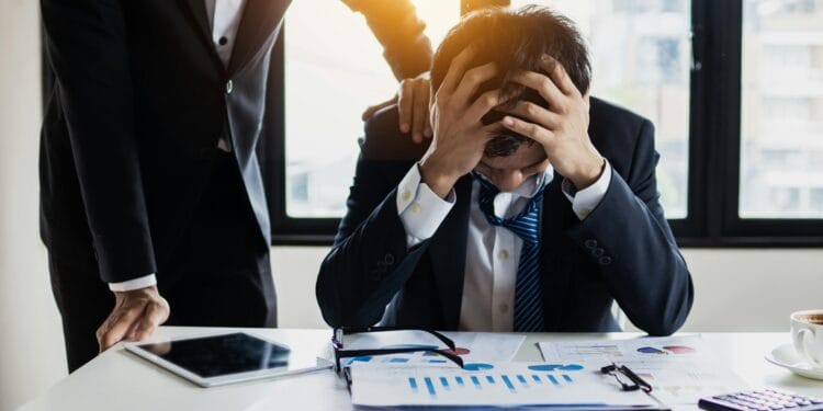 Professional feeling exhausted and wired under stressful workload. An overwhelmed businessman grasps his head, surrounded by reports in a bright office.