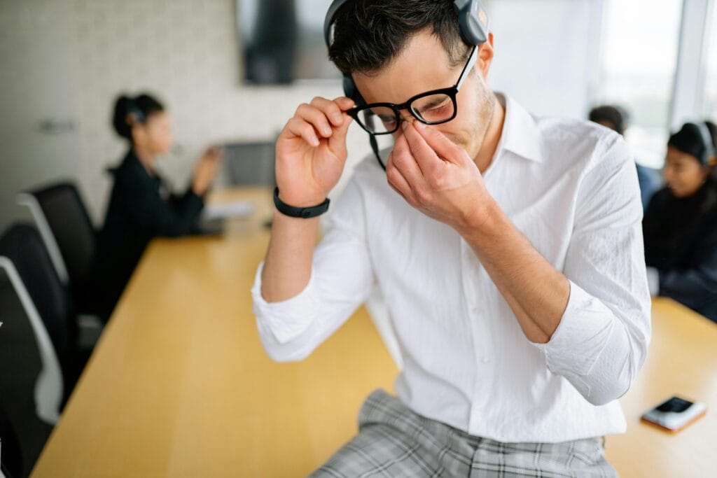 Young employee in white shirt sitting at office table, holding glasses and showing frustration during meeting. How to Overcome Emotional Burnout Naturally.