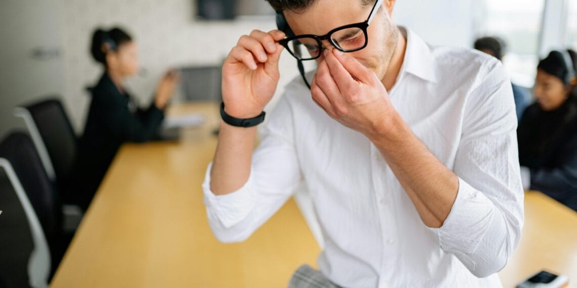 Young employee in white shirt sitting at office table, holding glasses and showing frustration during meeting. How to Overcome Emotional Burnout Naturally.