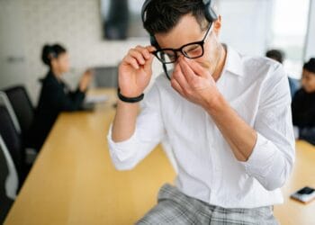 Young employee in white shirt sitting at office table, holding glasses and showing frustration during meeting. How to Overcome Emotional Burnout Naturally.