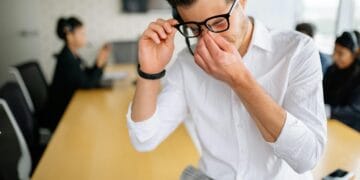 Young employee in white shirt sitting at office table, holding glasses and showing frustration during meeting. How to Overcome Emotional Burnout Naturally.