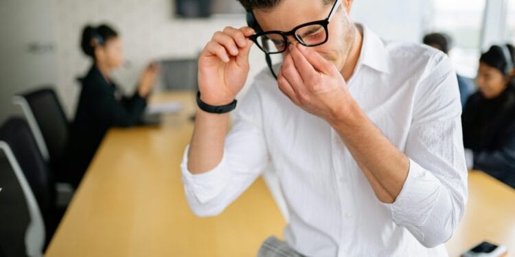 Young employee in white shirt sitting at office table, holding glasses and showing frustration during meeting. How to Overcome Emotional Burnout Naturally.