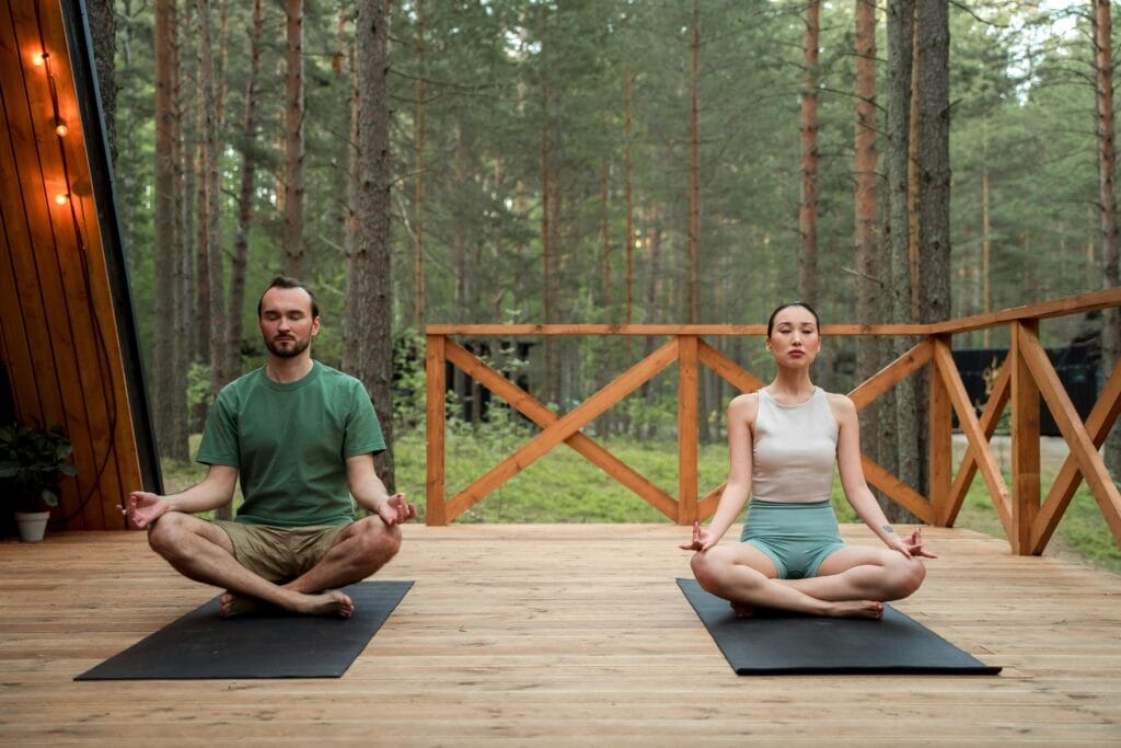 Man and woman meditating in lotus position on a wooden deck in a serene forest setting.