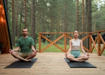 Man and woman meditating in lotus position on a wooden deck in a serene forest setting.