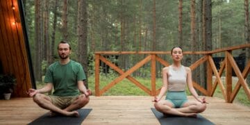 Man and woman meditating in lotus position on a wooden deck in a serene forest setting.