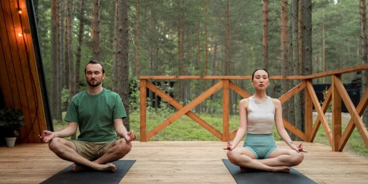 Man and woman meditating in lotus position on a wooden deck in a serene forest setting.